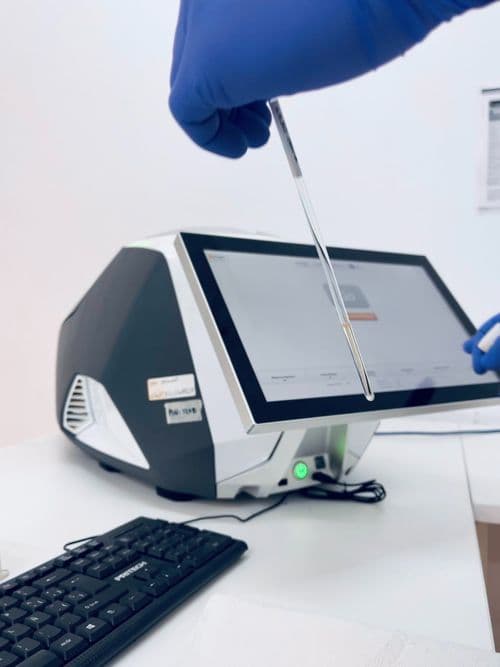 Una mano enguantada sosteniendo una herramienta de prueba sobre una máquina de análisis de laboratorio con pantalla táctil, con un teclado en primer plano.