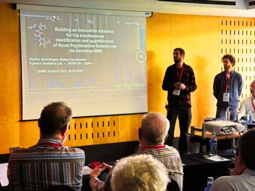 Un ponente de Kykeon Analytics hablando ante una audiencia en la Cumbre qNMR 2023, con la diapositiva de su presentación en pantalla.